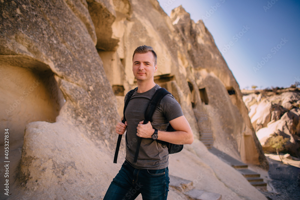 A young male tourist with a backpack stands on a background of rocks and looks at the camera.