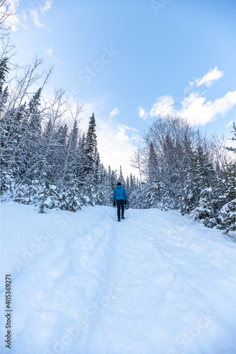 Man walking along a hiking trail in Canada with blue jacket and black jean pants in cold, freezing winter season surrounded by snow covered trees and boreal forest. 