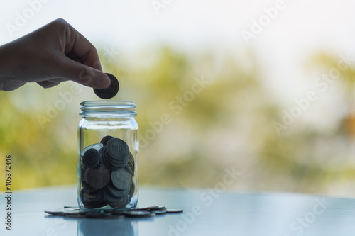 Closeup image of a hand collecting and putting coins in a glass jar for saving money concept
