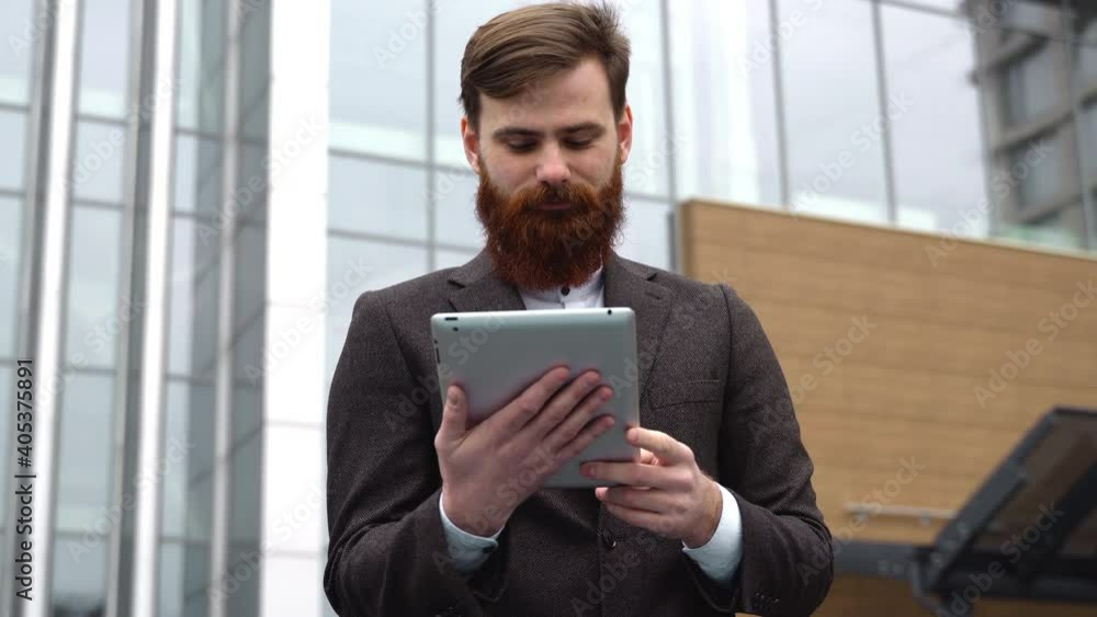 Modern Young bearded Business man working with a digital tablet. Young hipster businessman holding tablet in hands outdoor. Working online with a tablet while standing outside on an office building. Modern Young bearded Business man working with a digital tablet. Young hipster businessman holding tablet in hands outdoor. Working online with a tablet while standing outside on an office building.
