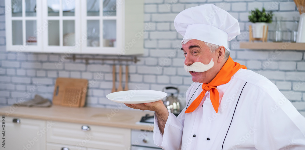 A friendly elderly chef is holding a plate in the kitchen. A gray-haired man with a thick gray mustache works in the kitchen A friendly elderly chef is holding a plate in the kitchen. A gray-haired man with a thick gray mustache works in the kitchen