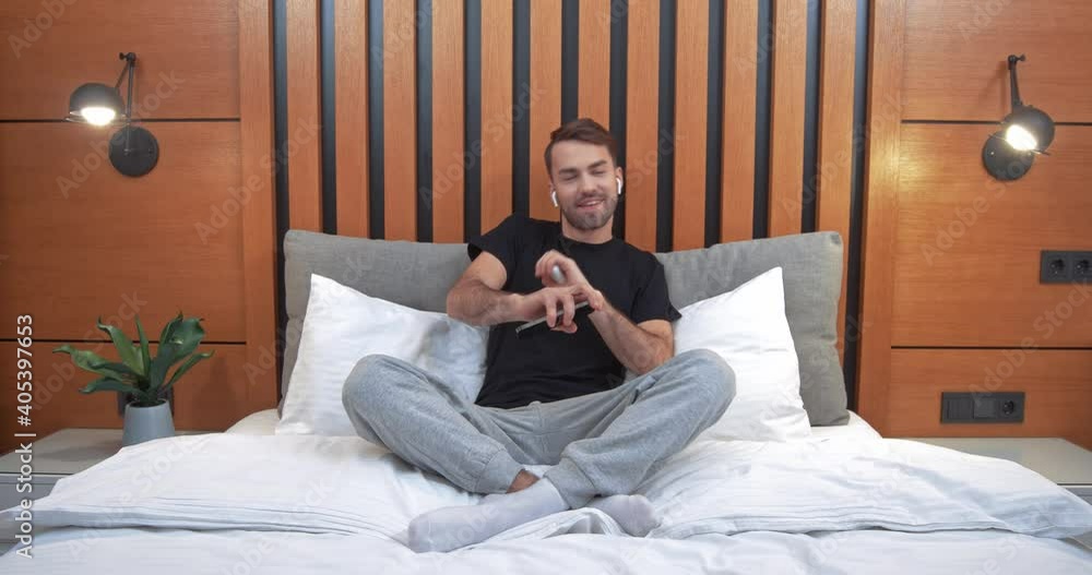 Happy man sitting on the bed in the bedroom and listening to music in wireless headphones, positive mood, music lover listens to music using a smartphone. Happy man sitting on the bed in the bedroom and listening to music in wireless headphones, positive mood, music lover listens to music using a smartphone.