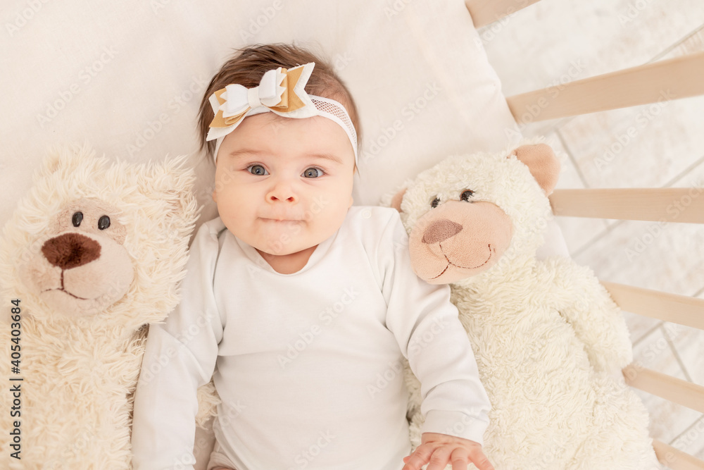 the baby is six months old in a crib in a white bodysuit with a Teddy bear