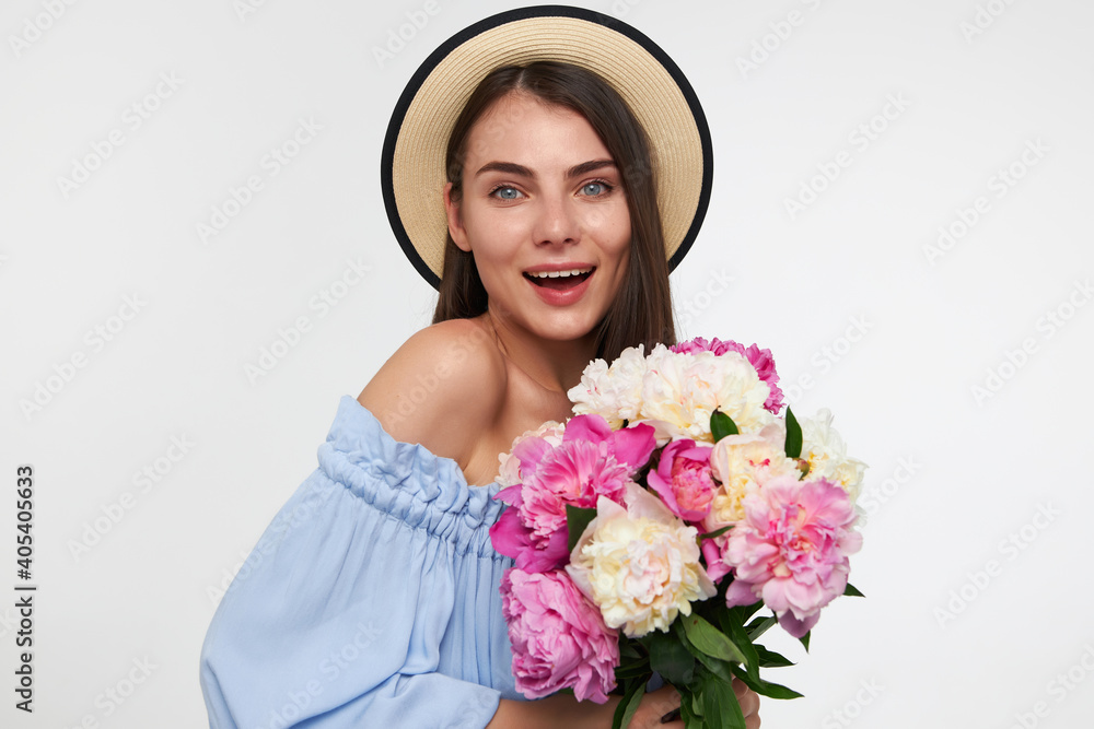 Portrait of attractive, nice looking girl with big smile and long brunette hair. Wearing a hat and blue dress. Holding bouquet of flowers and watching at the camera isolated over white background Portrait of attractive, nice looking girl with big smile and long brunette hair. Wearing a hat and blue dress. Holding bouquet of flowers and watching at the camera isolated over white background