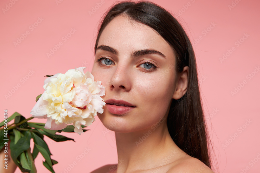 Young lady, pretty, charming woman with long brunette hair and healthy skin, touching cheek with a flower. Watching at the camera, closeup, isolated over pastel pink background Young lady, pretty, charming woman with long brunette hair and healthy skin, touching cheek with a flower. Watching at the camera, closeup, isolated over pastel pink background