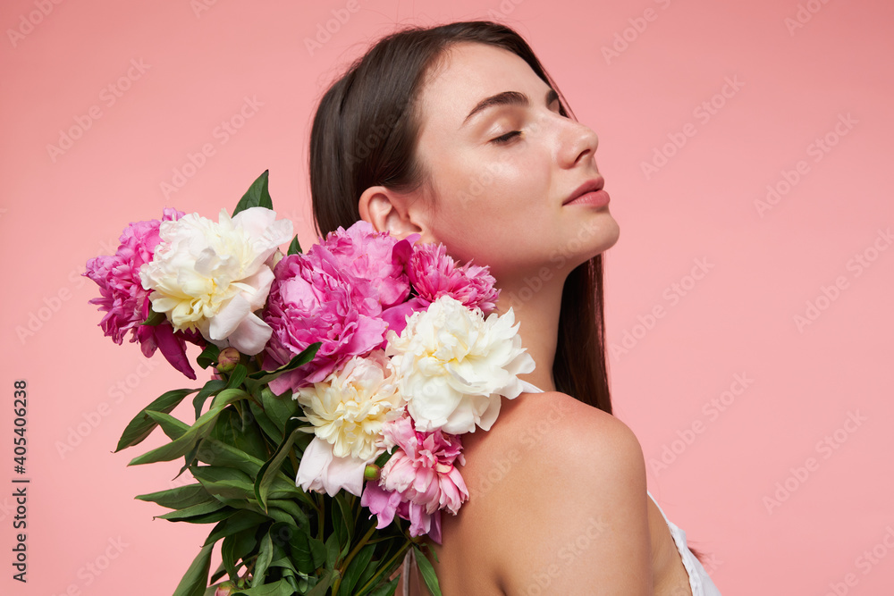 Nice looking woman, beautiful girl with long brunette hair, closed eyes and healthy skin. Wearing white dress and holds bouquet of flowers over here back. Stand isolated over pastel pink background