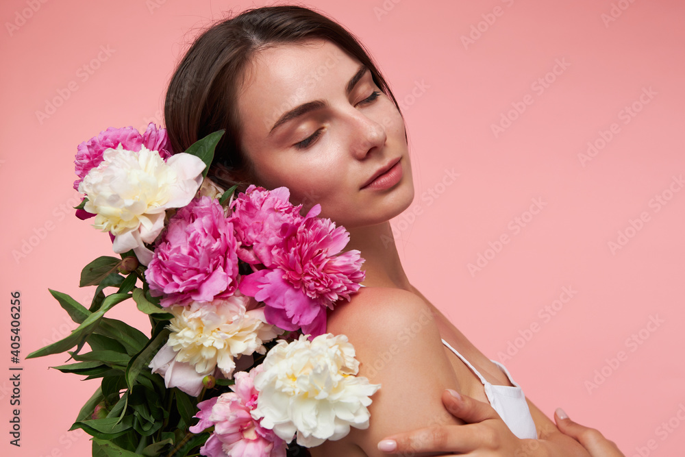 Portrait of attractive, nice looking girl with long brunette hair, closed eyes and healthy skin. Wearing white dress and holds bouquet of flowers. Stand isolated over pastel pink background