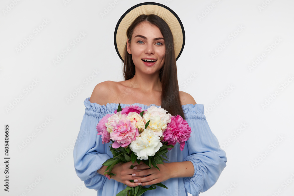 Portrait of attractive girl with big smile and long brunette hair. Wearing a hat and blue pretty dress. Holding a bouquet of beautiful flowers. Watching at the camera isolated over white background Portrait of attractive girl with big smile and long brunette hair. Wearing a hat and blue pretty dress. Holding a bouquet of beautiful flowers. Watching at the camera isolated over white background