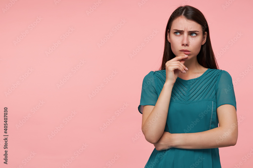 Teenage girl, serious looking woman with brunette long hair. Folding hands on a chest and touching her chin. Watching to the left at copy space over pastel pink background. Wearing emerald dress Teenage girl, serious looking woman with brunette long hair. Folding hands on a chest and touching her chin. Watching to the left at copy space over pastel pink background. Wearing emerald dress