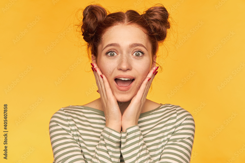 Teenage girl, happy looking red hair woman with two buns. Screams in surprise and touch cheeks, excited. Wearing striped sweater and watching at the camera isolated, closeup over yellow background Teenage girl, happy looking red hair woman with two buns. Screams in surprise and touch cheeks, excited. Wearing striped sweater and watching at the camera isolated, closeup over yellow background