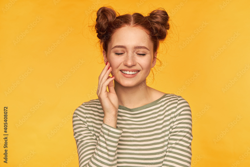 Portrait of attractive, charming red hair girl with two buns. Wearing striped sweater and smile with closed eyes, touching her cheek with fingertips. Stand isolated over yellow background Portrait of attractive, charming red hair girl with two buns. Wearing striped sweater and smile with closed eyes, touching her cheek with fingertips. Stand isolated over yellow background