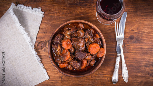 Ready burgundy beef in a clay bowl and a glass of red wine on a wooden table close-up, top view photo