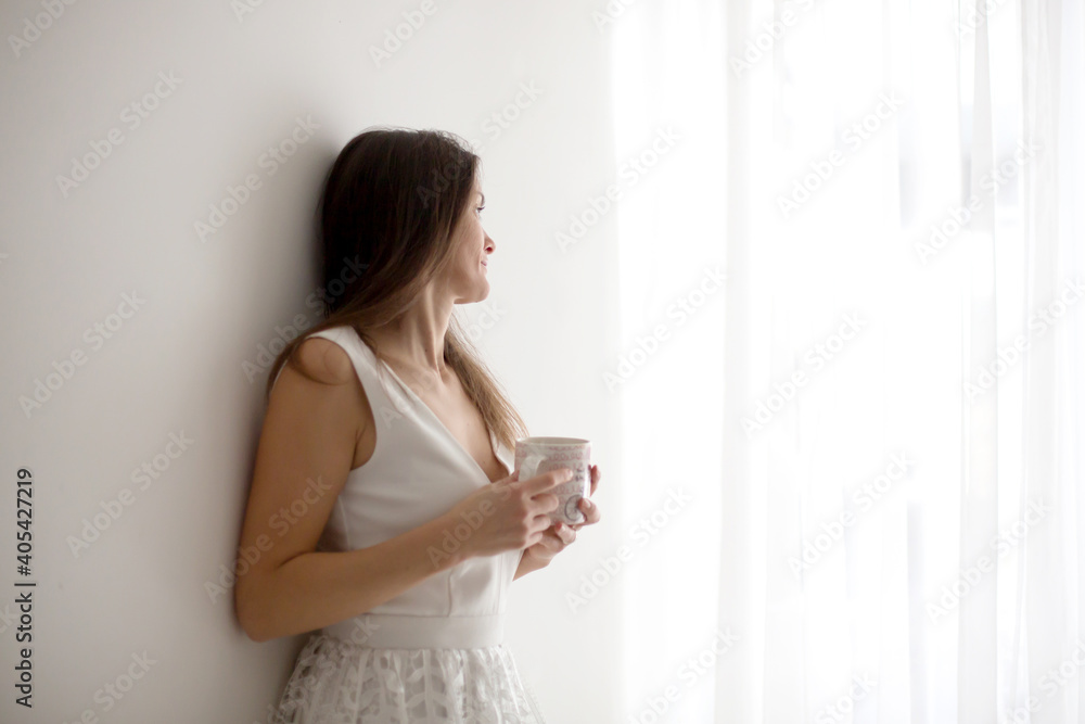 Middle age woman with white dress, drinking coffee at home, looking out of the window Middle age woman with white dress, drinking coffee at home, looking out of the window