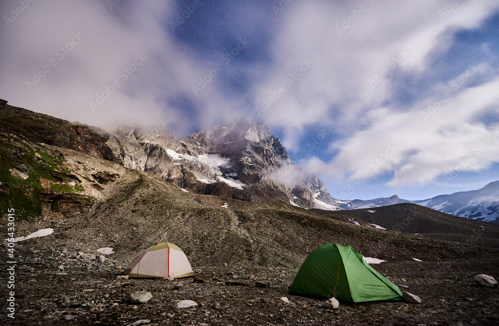 Panoramic view of night camping in night mountains. Scenery of tourist tents on rocky hill and mountain peak Matterhorn under beautiful misty sky with stars. Concept of travelling, hiking and camping.