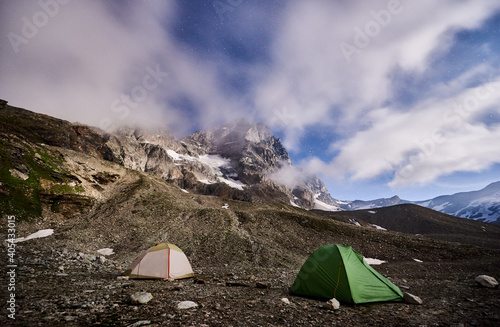 Panoramic view of night camping in night mountains. Scenery of tourist tents on rocky hill and mountain peak Matterhorn under beautiful misty sky with stars. Concept of travelling  hiking and camping.