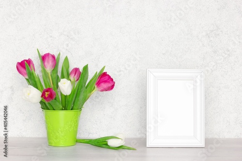 Fototapeta Naklejka Na Ścianę i Meble -  Blank white photo frame with tulip flower bouquet on the table near the wall. Selective focus. Spring mock-up background