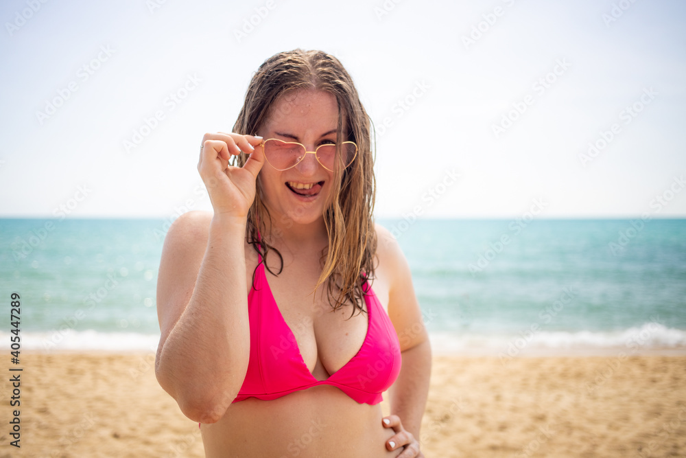 young playful blonde girl smiling, flirting and looking at the camera, dressed in a pink bikini and glasses with hearts.sea beach is on background. young playful blonde girl smiling, flirting and looking at the camera, dressed in a pink bikini and glasses with hearts.sea beach is on background.