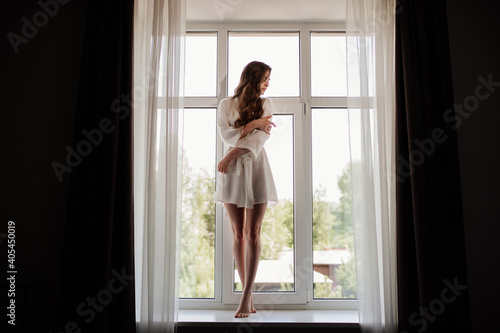 Beautiful sexy girl in a white robe with long hair stands in the window opening