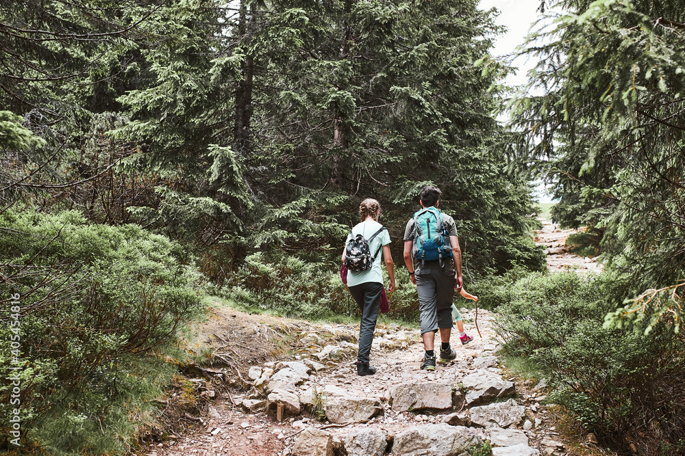 Family with backpacks hiking in a mountains actively spending summer vacation together walking on forest path talking and admiring nature mountain landscapes