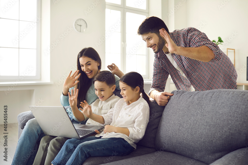 Staying at home and video calling relatives in lockdown during Covid-19 pandemic: Happy young family with little children sitting on sofa in living-room, smiling and waving hands at laptop computer Staying at home and video calling relatives in lockdown during Covid-19 pandemic: Happy young family with little children sitting on sofa in living-room, smiling and waving hands at laptop computer