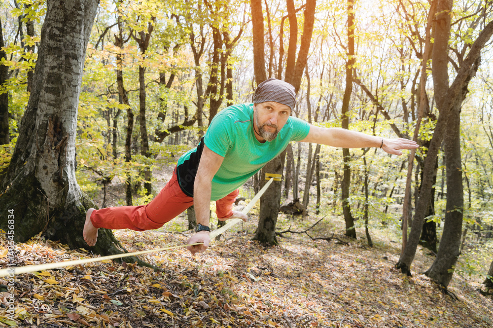 A bearded man in age balances while sitting on a taut slackline in the autumn forest. Outdoor Leisure