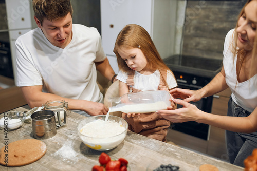 A full multicultural family with an adorable daughter gathered in a modern kitchen, preparing pancakes together. Making cake mix, pouring milk, socializing pleasure and culinary hobby
