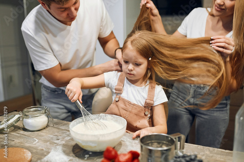 A full multicultural family with an adorable daughter gathered in a modern kitchen, preparing pancakes together. Making cake mix, stir up the dough, enjoy socializing and culinary hobbies