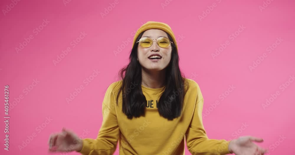 Portrait of young happy joyful Asian pretty female in yellow hat and glasses sending air kisses and making heart gesture while standing isolated on pink background and smiling. Positive mood concept Portrait of young happy joyful Asian pretty female in yellow hat and glasses sending air kisses and making heart gesture while standing isolated on pink background and smiling. Positive mood concept