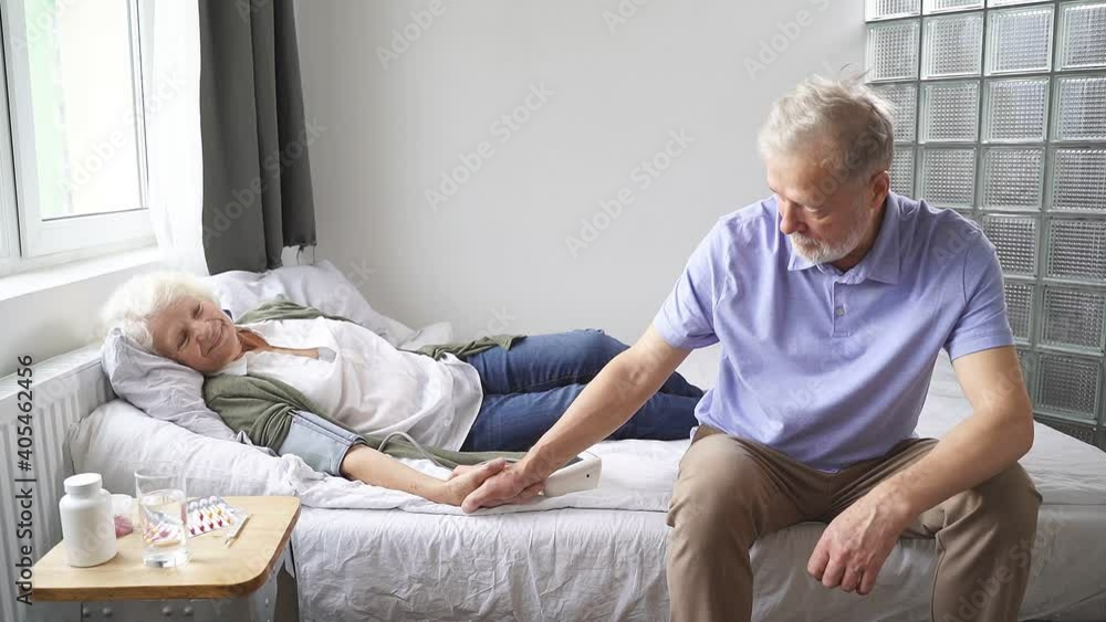 elderly woman is lying on a bed and is ill, and her husband is sitting next to her and supporting his wife by holding her hand. The concept of happiness together. elderly woman is lying on a bed and is ill, and her husband is sitting next to her and supporting his wife by holding her hand. The concept of happiness together.