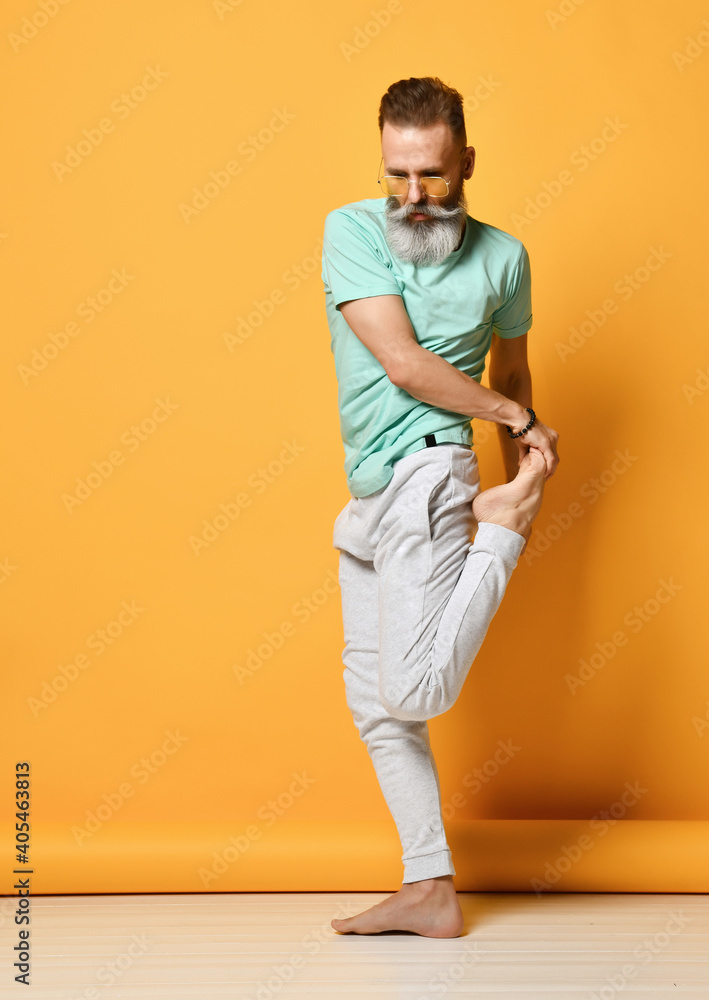 Mature handsome grey-haired brutal man wearing trendy sportswear stretching leg standing in yoga position, practicing asana exercise and workout on balance. Studio shot on yellow background Mature handsome grey-haired brutal man wearing trendy sportswear stretching leg standing in yoga position, practicing asana exercise and workout on balance. Studio shot on yellow background