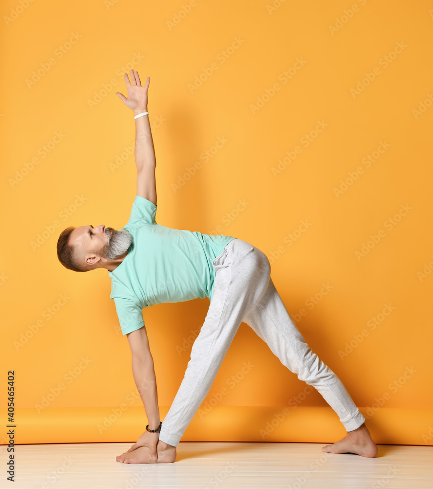 Mature handsome bearded gray-haired man in sportswear practicing yoga, standing in the pose of an elongated triangle Utthita Trikonasana. Sport, healthy lifestyle and care. Full-length indoor studio
