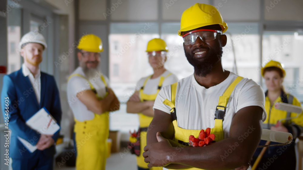 Portrait of afro-american painter in uniform holding roller brush posing with team on background Portrait of afro-american painter in uniform holding roller brush posing with team on background