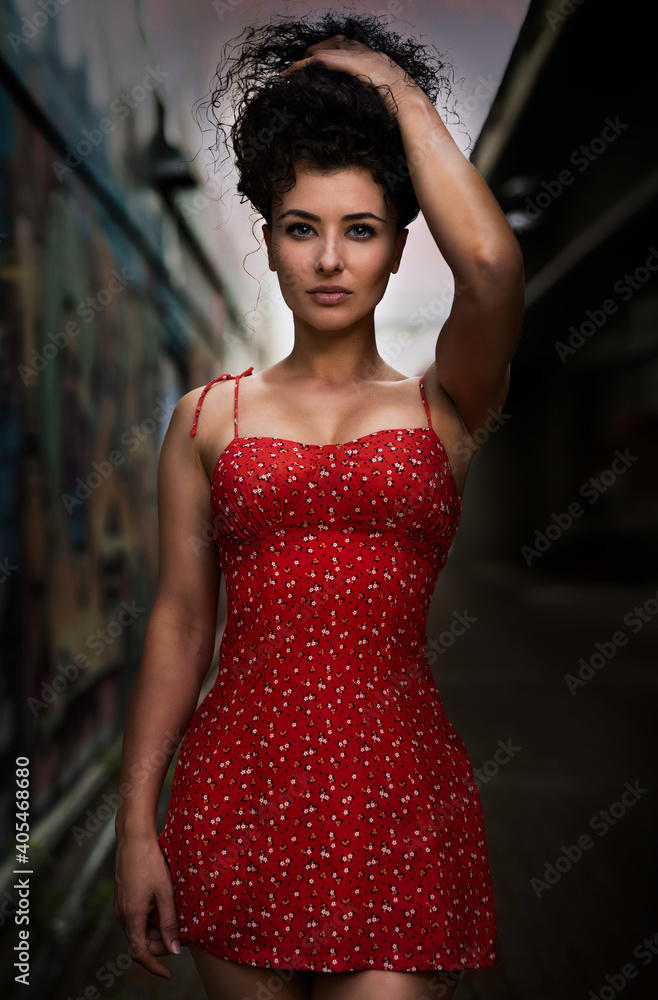 Fototapeta premium A beautiful young curly-hair woman in a little red dress is walking on the street. - Portrait Photography. 