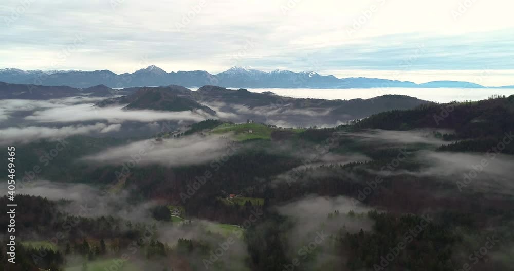 Drone flying over hills covered with forest and low clouds in Slovenia. Aerial view of Saint Thomas church and Alps mountains in the distance. Elevated view of amazing landscape. Slow descending Drone flying over hills covered with forest and low clouds in Slovenia. Aerial view of Saint Thomas church and Alps mountains in the distance. Elevated view of amazing landscape. Slow descending