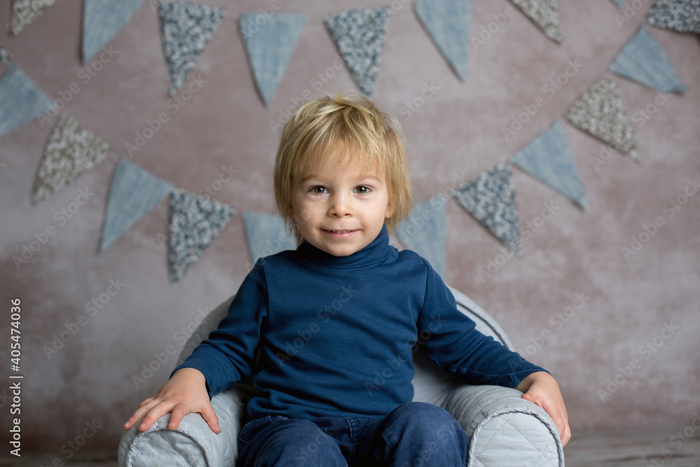 Fototapeta premium Cute blond toddler child, boy, sitting on little baby armchair, reading a book and holding flowers