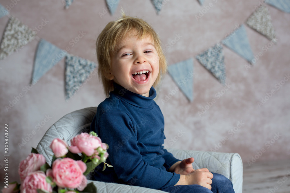 Fototapeta premium Cute blond toddler child, boy, sitting on little baby armchair, reading a book and holding flowers