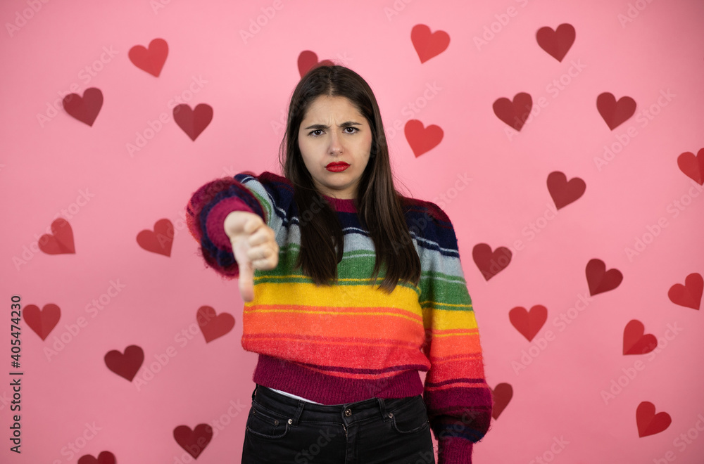 Young beautiful woman over pink background with harts with angry face, negative sign showing dislike with thumb down Young beautiful woman over pink background with harts with angry face, negative sign showing dislike with thumb down
