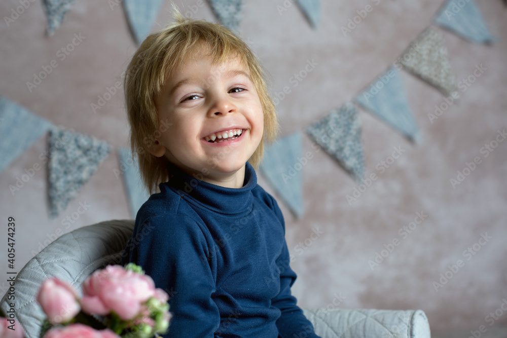 Cute blond toddler child, boy, sitting on little baby armchair, reading a book and holding flowers Cute blond toddler child, boy, sitting on little baby armchair, reading a book and holding flowers