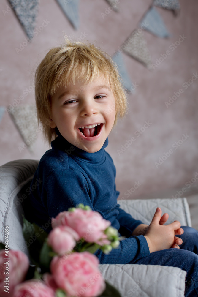 Fototapeta premium Cute blond toddler child, boy, sitting on little baby armchair, reading a book and holding flowers