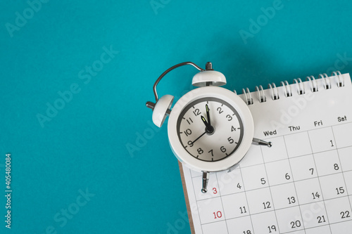 white analog alarm clock on blurred calendar with grunge blue background with copy space, business meeting schedule, travel planning or project milestone and reminder   concept ,panorama