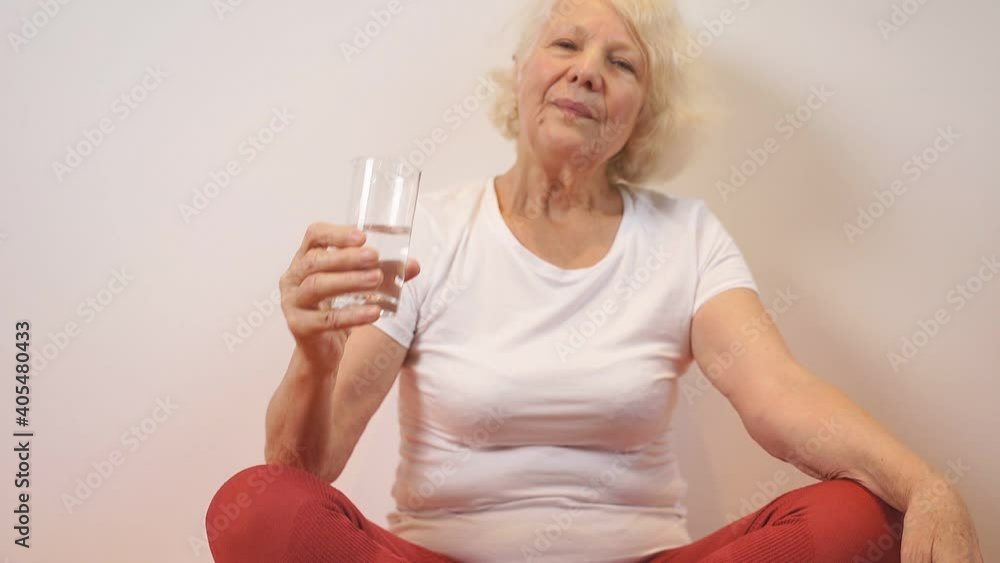 positive woman of retirement age drinks water after yoga and fitness classes, sitting at home on a mat. positive woman of retirement age drinks water after yoga and fitness classes, sitting at home on a mat.