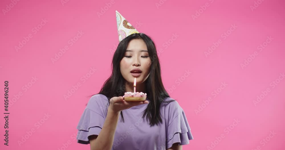 Close up portrait of joyful Asian pretty young woman in purple blouse in b-day hat blowing candle on birthday biscuit mini cake while standing on pink background celebrating making a wish Close up portrait of joyful Asian pretty young woman in purple blouse in b-day hat blowing candle on birthday biscuit mini cake while standing on pink background celebrating making a wish