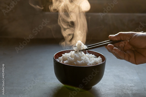 The man hand of using black chopsticks holding hot jasmine rice with smoke and steam in black bowl over dark background.hot food concept