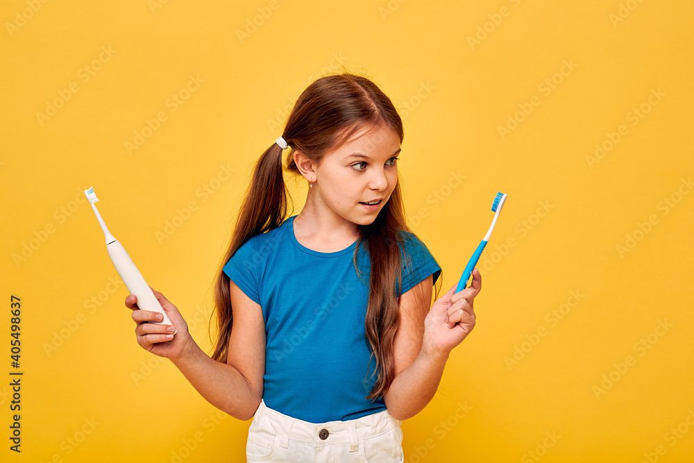 Pretty little girl holds manual and electric sonic toothbrush. Child selects of kind of toothbrush, on yellow background Pretty little girl holds manual and electric sonic toothbrush. Child selects of kind of toothbrush, on yellow background