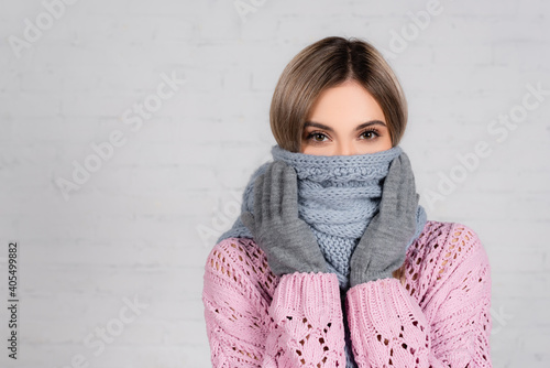 Woman in knitted sweater and gloves wrapped in scarf looking at camera on white background