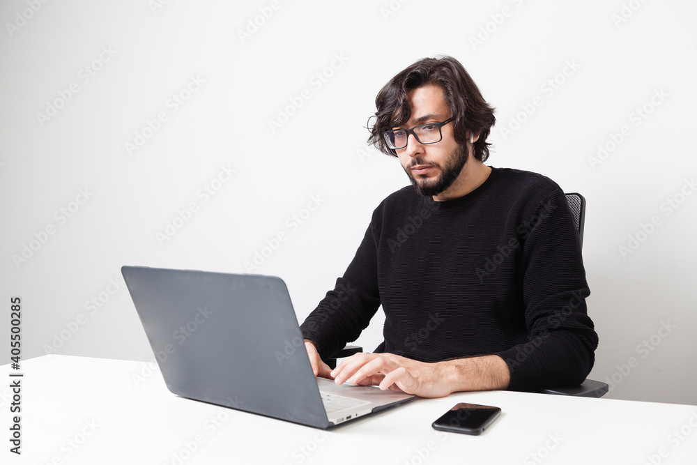 Middle eastern adult bearded man with long hair and glasses using laptop computer with smartphone on a white office room with copy space. Remote working concept. Middle eastern adult bearded man with long hair and glasses using laptop computer with smartphone on a white office room with copy space. Remote working concept.
