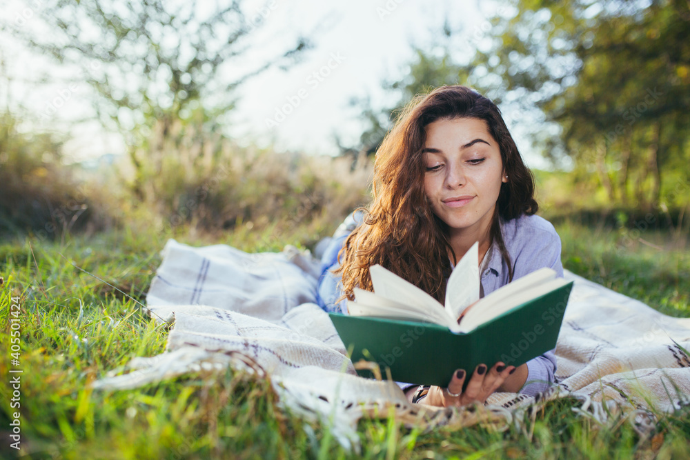 Beautiful young woman with curly black hair, resting lying on a mat in the park on the grass, and holding a book reads Beautiful young woman with curly black hair, resting lying on a mat in the park on the grass, and holding a book reads