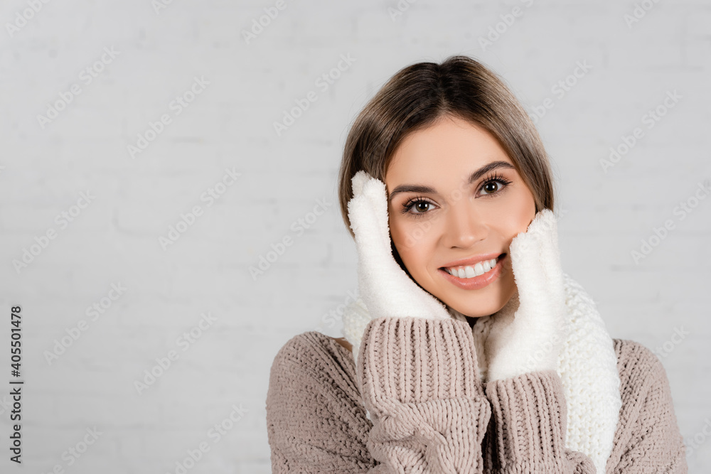 Young woman in sweater, knitted scarf and gloves with hands near face smiling at camera on white background Young woman in sweater, knitted scarf and gloves with hands near face smiling at camera on white background