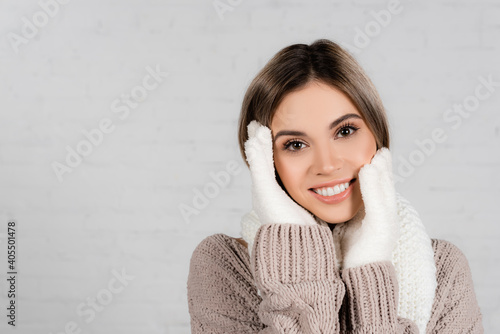 Young woman in sweater, knitted scarf and gloves with hands near face smiling at camera on white background