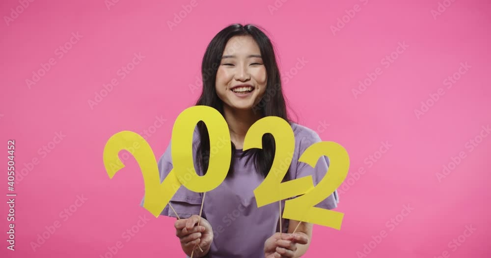 Portrait of cheerful happy Asian young positive female teen smiling and looking at camera standing in studio isolated on pink rosy background holding 2022 sign in hands celebrating New Year.
